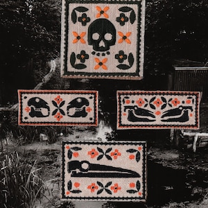 May include: Four embroidered patches with a Day of the Dead theme. The patches feature black skulls and other motifs, with orange and black floral accents on a beige background. The patches are framed with a dark green border.