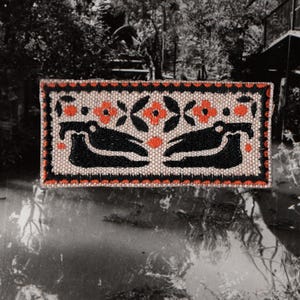 May include: Embroidered rectangular patch with a beige background, featuring black bird-like figures and orange floral designs. The border is black and orange. The patch is set against a monochrome outdoor scene.