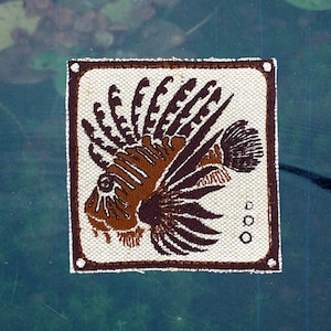 Puede incluir: Textil cuadrado blanco roto con un diseño de pez león marrón y marrón oscuro. El pez león está centrado, con aletas detalladas y un aspecto texturizado. El textil está bordeado por un marco marrón, sobre un fondo de agua azul-verdosa.