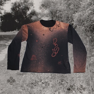 Könnte beinhalten: Schwarzes Langarmshirt mit Rundhalsausschnitt und Farbverlauf. Das Oberteil hat ein braunes und schwarzes Farbschema mit roten abstrakten Formen und einem gesprenkelten Muster. Die Ärmel sind einfarbig schwarz.