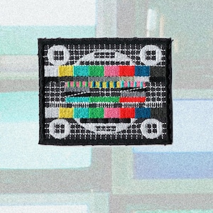Könnte beinhalten: Gestickter Aufnäher mit einem klassischen Fernseh-Testbild-Design. Der Aufnäher hat einen schwarzen Rand und einen Hintergrund mit Gittermuster. Das Testbild enthält farbige horizontale Balken und weiße Kreise, die eine Retro-Ästhetik erzeugen.