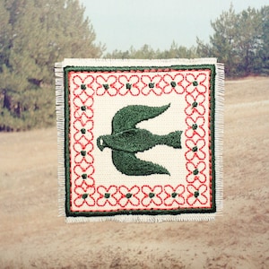 Op de afbeelding: Een vierkante stoffen patch met een crèmekleurige basis en een centraal geborduurde donkergroene vogel. Het ontwerp is omlijst door een rand van rode geborduurde bloemen met donkergroene harten, afgewerkt met een rafelige, gefranjerde rand.