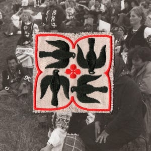 Puede incluir: Textil cuadrado bordado con un borde rojo, con cuatro diseños de pájaros negros y una flor roja en el centro. El fondo muestra una multitud de personas, lo que sugiere un evento cultural o una reunión.