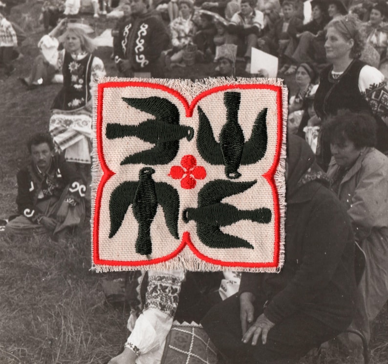 Puede incluir: Textil cuadrado bordado con un borde rojo, con cuatro dise&ntilde;os de p&aacute;jaros negros y una flor roja en el centro. El fondo muestra una multitud de personas, lo que sugiere un evento cultural o una reuni&oacute;n.