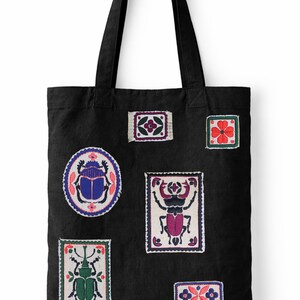 May include: Black tote bag featuring embroidered patches with insect and floral designs. The patches are in various shapes and sizes, with colors including blue, green, red, and purple. The bag has two black handles.