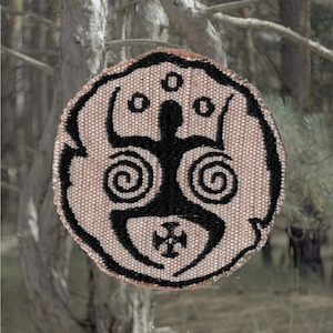 Puede incluir: Parche circular con un fondo tejido beige y un diseño tribal negro. El diseño presenta una figura humana estilizada con espirales y una cruz. El parche tiene un borde negro.