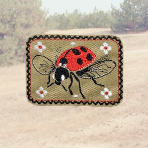 Peut inclure: Écusson brodé représentant une coccinelle rouge à pois noirs et ailes, sur fond beige. Le patch a une bordure en zigzag noire et rouge et de petits accents de fleurs blanches. Le design est rectangulaire.