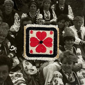 May include: A square, embroidered textile with a red floral design on a beige background, framed by a black border. The image is set against a backdrop of a group of people in traditional clothing.