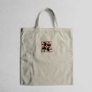 Puede incluir: Bolso tote beige con un dise&ntilde;o cuadrado en el centro. El dise&ntilde;o presenta formas negras con forma de p&aacute;jaro y formas verdes con forma de hoja, delineadas en rojo. El bolso tiene un asa simple y est&aacute; hecho de tela clara.