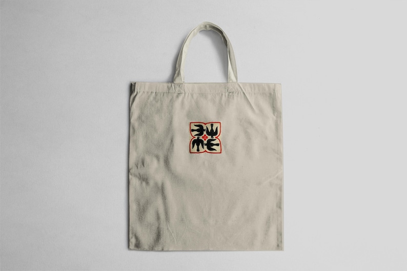 Puede incluir: Bolso tote beige con un dise&ntilde;o cuadrado en el centro. El dise&ntilde;o presenta formas negras con forma de p&aacute;jaro y formas verdes con forma de hoja, delineadas en rojo. El bolso tiene un asa simple y est&aacute; hecho de tela clara.