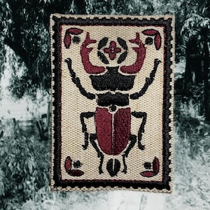 Op de afbeelding: Een rechthoekige beige stoffen patch met een zwart en bordeaux scarabee-ontwerp. De kever heeft een bordeauxrood lichaam en hoofd, met zwarte poten en antennes. De patch is omlijst door een zwarte rand.