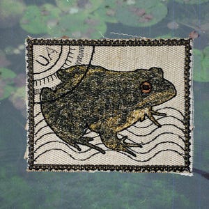 May include: Embroidered patch with a frog design. The frog is in green and gold, with a detailed outline and a reddish-brown eye. The patch is rectangular with a black border and a cream background. The design is reminiscent of a postage stamp.