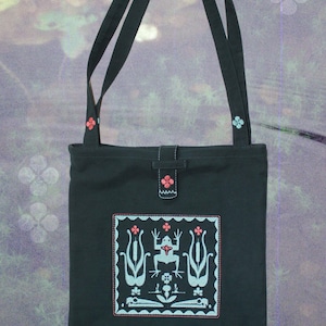 Könnte beinhalten: Schwarze Tragetasche mit langen Trägern und einem quadratischen Stickdesign. Das Design zeigt einen Frosch und florale Elemente in Hellblau und Rosa. Die Tasche hat einen Knopfverschluss und florale Akzente.