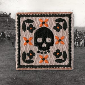 Puede incluir: Parche de tela cuadrado con una calavera negra, flores naranjas en forma de cruz y diseños florales negros. El parche tiene un fondo beige y un borde negro con un borde marrón. El diseño está bordado.