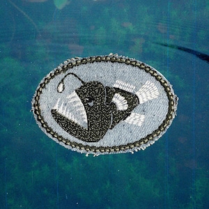 May include: An oval-shaped patch featuring a black anglerfish design with white accents. The patch is embroidered on a light blue denim-like fabric and has a decorative border. The anglerfish has a prominent lure.