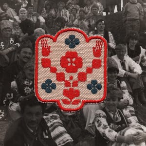 May include: A vintage embroidered patch with a red and blue floral design. The patch has a beige background and a red border. The design includes a central flower, four blue flowers, and two red hand motifs. The background is a black and white photo of a group of people.