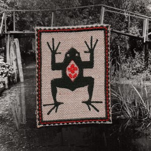 May include: A rectangular patch with a frog design. The frog is black with a red floral design on its belly. The patch has a tan background with a red border. The frog is in a symmetrical pose with its arms and legs spread out.