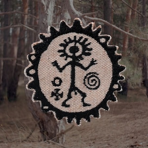 Może przedstawiać: Okrągła, tkana naszywka z czarnym wzorem postaci plemiennej. Postać znajduje się na beżowym tle, otoczona czarną, karbowaną obwódką. Naszywka przedstawia stylizowaną postać ludzką, spiralę i inne symbole.