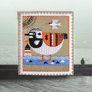May include: A rectangular embroidered patch on a tan fabric background featuring a stylised bird with black, white, and orange plumage. The design includes a small lighthouse, a flying bird, and the text Norway. The border is white and serrated.