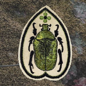 Könnte beinhalten: Gestickter Aufnäher mit einem grünen Käferdesign. Der Käfer ist mit schwarzen Beinen und Fühlern versehen, vor einem cremefarbenen Hintergrund mit schwarzem Rand. Oben befindet sich ein grünes Blumenmuster.