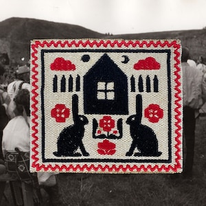 Può includere: Una toppa quadrata ricamata con un design di arte popolare. Presenta una casa nera, due conigli neri, fiori rossi e nuvole rosse su sfondo crema. Il bordo è rosso con un motivo a zig-zag.