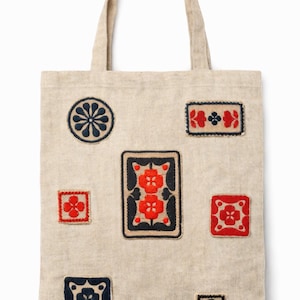 May include: Natural-coloured tote bag adorned with embroidered floral and geometric motifs in red and navy blue. The bag showcases various shapes, including circles, squares, and rectangles, each with a unique pattern. It has a simple, robust design with a handle.