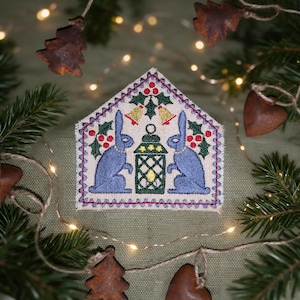 Pode incluir: Um ornamento de tecido em forma de casa com bordado de dois coelhos azuis voltados para uma lanterna verde central. O design inclui bagas de azevinho vermelhas, folhas verdes e dois pequenos sinos amarelos, contornado por uma costura azul.