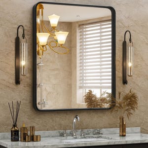 May include: A rectangular black-framed mirror hangs above a marble countertop in a bathroom setting. The mirror reflects a window with blinds and a gold chandelier with three lights. Two black sconces with cylindrical glass shades flank the mirror.