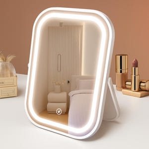 May include: A white, rectangular vanity mirror with a bright LED light border. The mirror reflects a bedroom scene with a bed, nightstand, and decorative wall panel. Makeup products, including lipstick and foundation, are displayed on a wooden block.