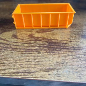 Peut inclure: Une petite benne à ordures en plastique orange de forme rectangulaire avec des barreaux verticaux sur les côtés.