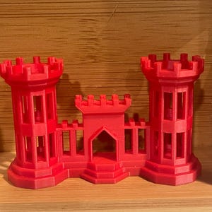 May include: A red 3D printed castle with two towers and a gate. The castle is made of plastic and has a detailed design.