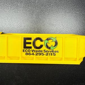 May include: A bright yellow miniature dumpster model featuring vertical structural ribs along the side. The text ECO with a green recycling symbol, followed by ECO Waste Services and the phone number 864-295-2115, is printed in black on the side.