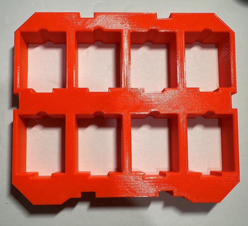 May include: A bright orange plastic storage case with nine rectangular compartments. The case is designed to hold small items, such as game cards or other collectibles.