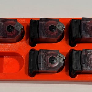 May include: An orange plastic tray holds five black and clear magazines. The magazines are arranged in a rectangular pattern, with a space for a sixth magazine. The tray is a bright, solid orange color.