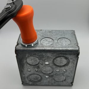 May include: A close-up of an orange plastic electrical box cover being installed with pliers. The cover has a round shape and is being pressed into a metal electrical box with multiple holes.