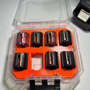May include: A clear plastic storage case with an orange interior holds six black and red router bits. The case has a black latch on the front.