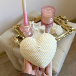 May include: A white heart-shaped candle with a honeycomb pattern. The candle is on a gold tray with a pink candle holder and a white rose.