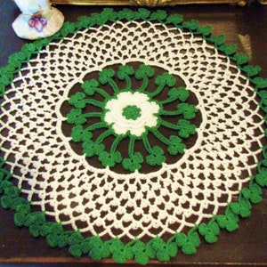 May include: A white crocheted doily with a green shamrock design around the edge. The doily is on a dark brown surface.