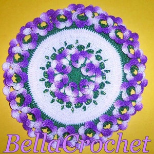 Op de afbeelding: Een gehaakt tafellaken met een wit midden en een groene en paarse rand. Het tafellaken is versierd met paarse en witte bloemen.