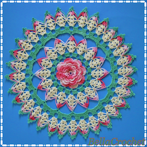 May include: A crocheted doily with a floral design in shades of pink, green, and white. The doily has a central flower with multiple layers of petals.