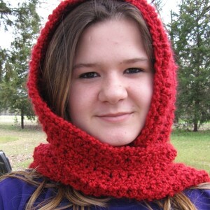 May include: A young person wearing a red crocheted hooded scarf. The scarf is thick and has a textured pattern. The person is wearing a purple shirt.