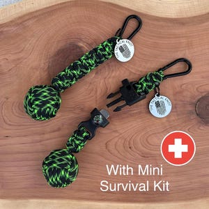 May include: Two black and green paracord survival bracelets with a small compass and whistle attached. Each bracelet has a silver charm with the text "The Tactical Knot" and an American flag design. The bracelets are on a wooden surface with a red cross symbol and the text "With Mini Survival Kit".