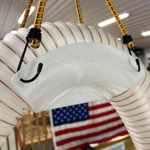 May include: A white, curved object with a semi-transparent, textured surface, suspended by black hooks and yellow and red bungee cords. The object has a ribbed design with copper-colored lines. An American flag is visible in the background.