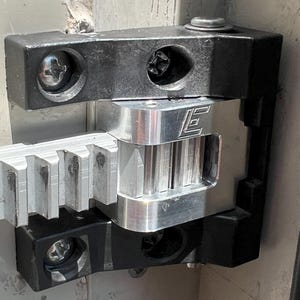 Op de afbeelding: Close-up van een mechanische constructie met een zilverkleurige tandheugel en een zilver-zwarte metalen component. De component is bevestigd met schroeven en heeft de letter "E" gegraveerd. De constructie maakt waarschijnlijk deel uit van een grotere machine.