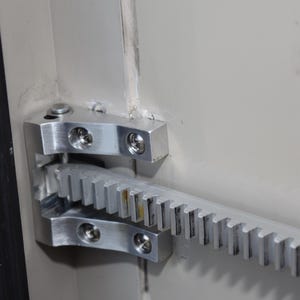 May include: Close-up of a silver metal door hinge with a gear rack attached to the door frame.
