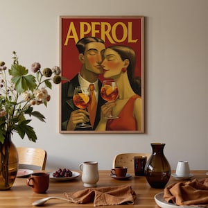 May include: A framed vintage-style poster with the word "APEROL" in yellow lettering. The artwork depicts a couple toasting with Aperol cocktails. The man wears a suit and orange tie, and the woman wears a red dress. The poster is displayed on a wooden table with various dining accessories.