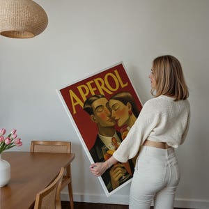 May include: A framed Aperol poster featuring a couple kissing, with the word "APEROL" in yellow letters on a red background. The poster is held by a person wearing a white sweater and pants.