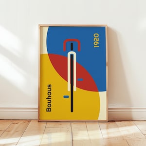 Può includere: Una stampa artistica Bauhaus incorniciata con un design geometrico che ricorda una bicicletta. L'opera utilizza colori primari: blu, rosso e giallo, con il testo "Bauhaus" e "1920". La stampa è esposta su un pavimento in legno.