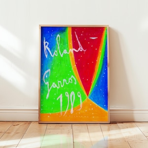 Puede incluir: Una obra de arte enmarcada con un diseño abstracto colorido. La obra presenta una vibrante mezcla de tonos azules, verdes, rojos y naranjas. El texto blanco en cursiva dice "Roland Garros 1989". La obra se exhibe sobre un suelo de madera contra una pared blanca.