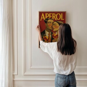 May include: A framed art print featuring a couple holding cocktails, with the word "APEROL" in yellow. The artwork has a vintage style with a red background and a brown frame. The print is being hung on a white wall.